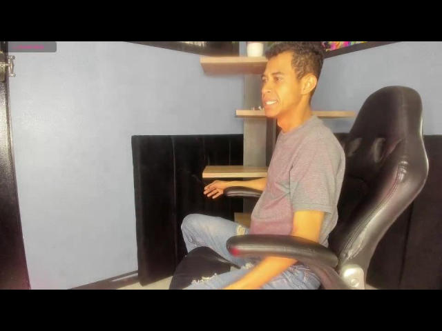 ZarekBlack, sitting in a black office chair, wearing a grey t-shirt and blue jeans, smiling at the camera, with a small wooden desk to his right.
