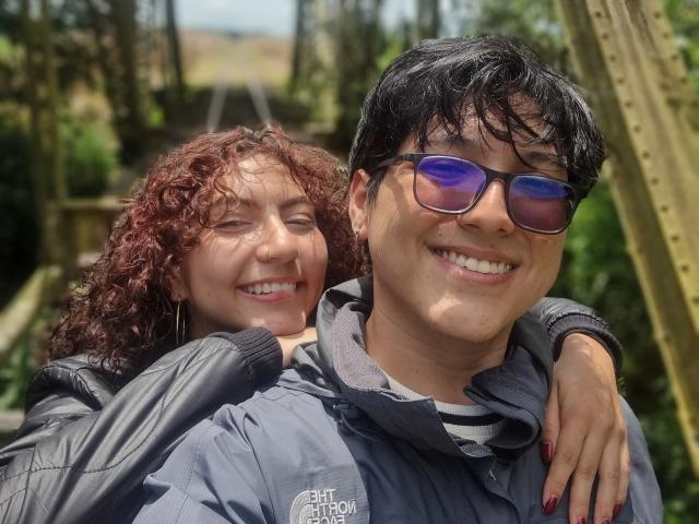 VanillaHotG with curly red hair and small tits hugs VanillaHotG with black hair, blue sunglasses, and a smile, wearing a grey jacket. Outside on a sunny day.
