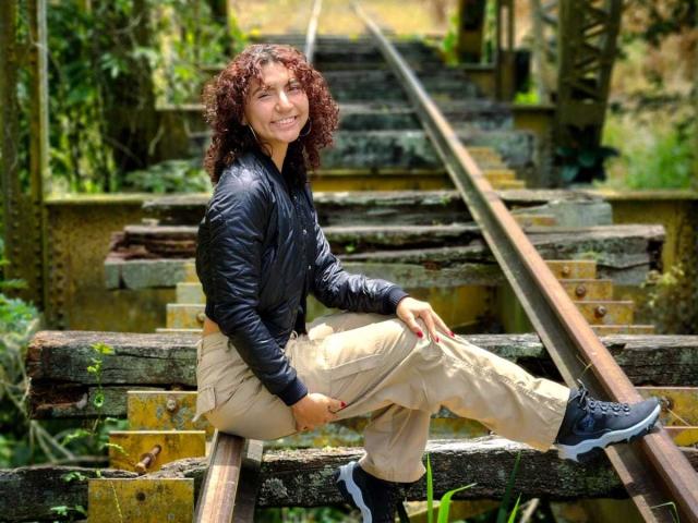 VanillaHotG with curly brown hair, black jacket, beige pants, and blue sneakers sits on rusty train tracks, smiling. Her ass is partially covered by her pants.
