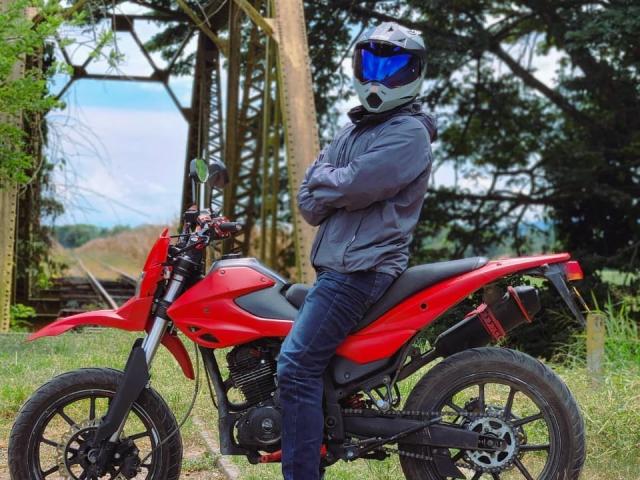 VanillaHotG in a gray jacket and blue jeans, wearing a black helmet with a blue visor, standing with arms crossed on a red motorcycle in a grassy area.