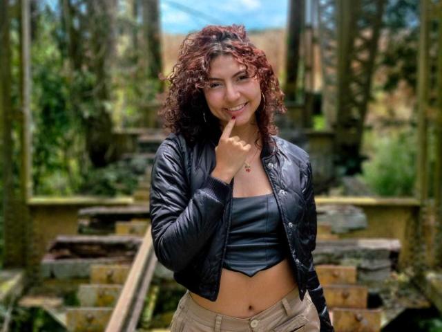 VanillaHotG with curly red-brown hair, black leather crop top, black jacket, and beige pants, finger on lips, smiling, standing in a wooded, ruined staircase area.