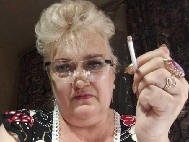 OneDamaX has grey curly hair, glasses, and is holding a cigarette with a pink tip, wearing floral and lace-patterned clothing, with multiple rings on her fingers.