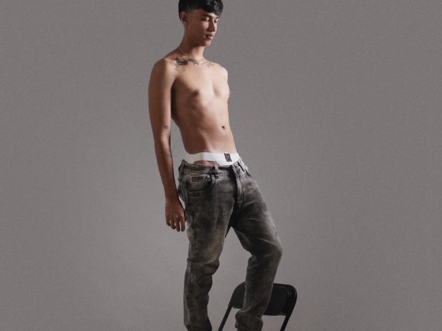 AtlasCody está sem camisa, vestindo jeans cinzas, levemente desbotados, com uma faixa branca na cintura, em pé ao lado de uma cadeira preta, olhando para baixo com um leve sorriso, com tatuagens no peito..