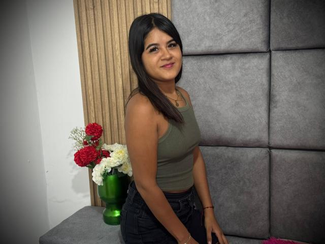 NaylaDiamond with medium tits, olive skin, dark straight hair, green tank top, black jeans, small red and white flower vase, gray padded wall, smiling, standing, brown wooden panel.