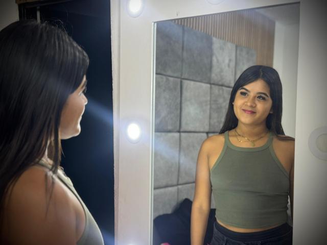 NaylaDiamond with long black hair, green crop top, and necklace, smiling at her reflection in a mirror with round bulbs, showing her small tits and cute face.