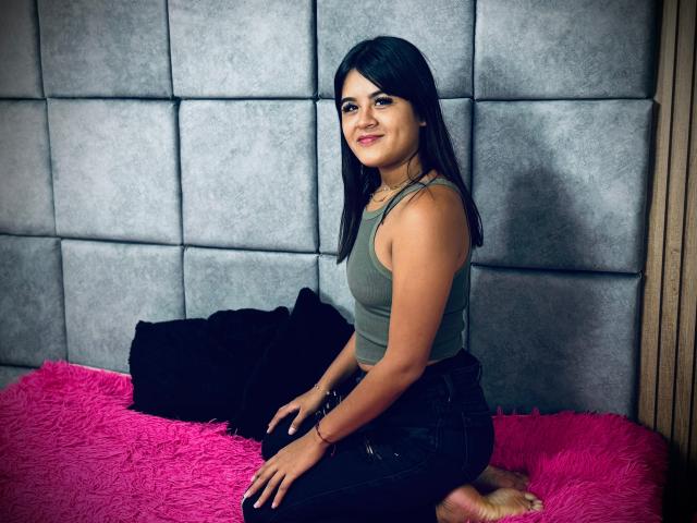 NaylaDiamond with dark hair, green tank top, and black pants, sitting on a pink fuzzy bed, smiling, showing her small ass and cute face, against a gray wall.