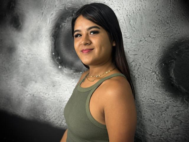 NaylaDiamond with medium brown skin, dark straight hair, green tank top, gold necklace, smiling slightly, standing against textured gray wall, showing her tits and a subtle ass curve.