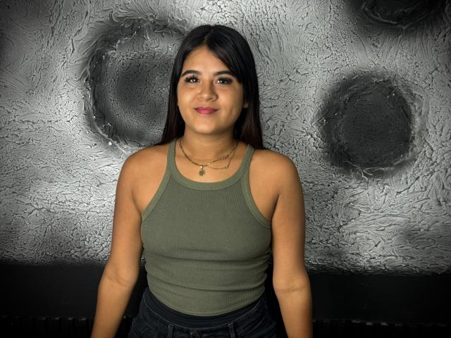 NaylaDiamond, with medium brown skin and long black hair, wears a green tank top and dark jeans, smiling at the camera, showing her small tits and a gold necklace.