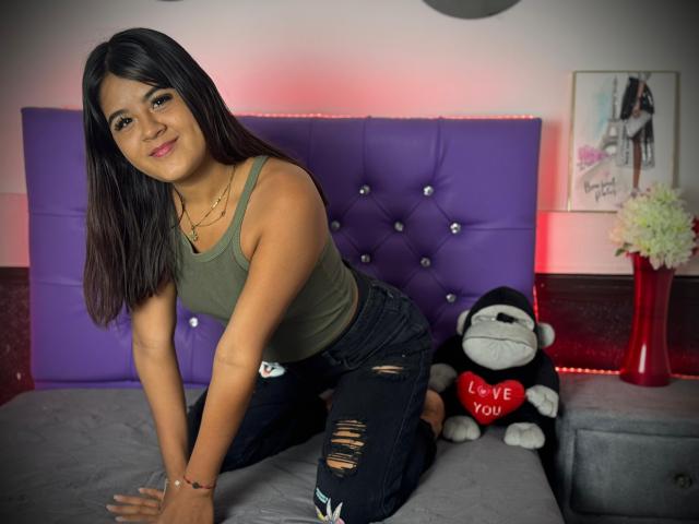 NaylaDiamond with dark brown hair, green tank top, and ripped black jeans, kneeling on a purple tufted bed, smiling, wearing a bracelet, with a "Love You" monkey toy.