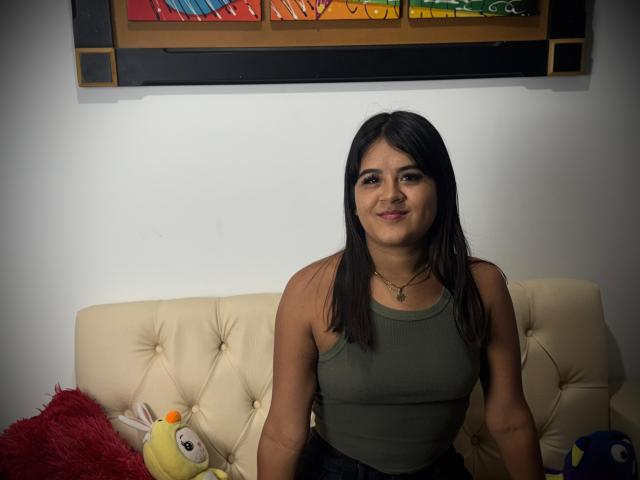 NaylaDiamond, a curvy woman with medium tits, sits on a cream couch, wearing a green tank top, black pants, and a necklace, smiling at the camera.