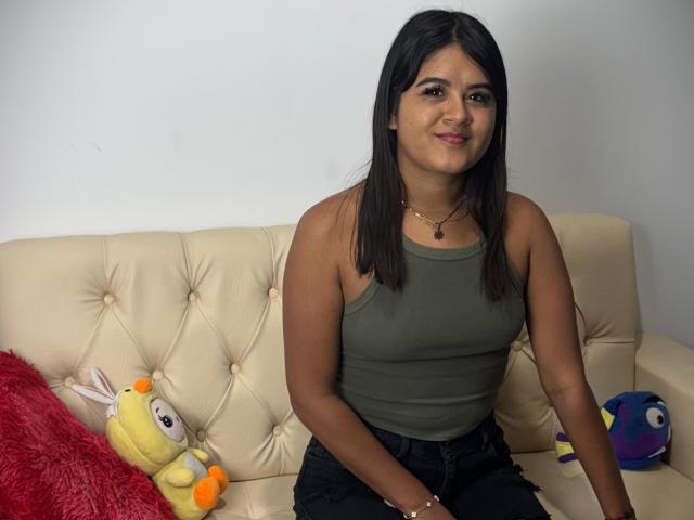 NaylaDiamond, dark-haired woman in green tank top, sitting on cream couch, black pants, red pillow, yellow bunny plushie, blue fish plushie, slight smile, necklace, tan skin.