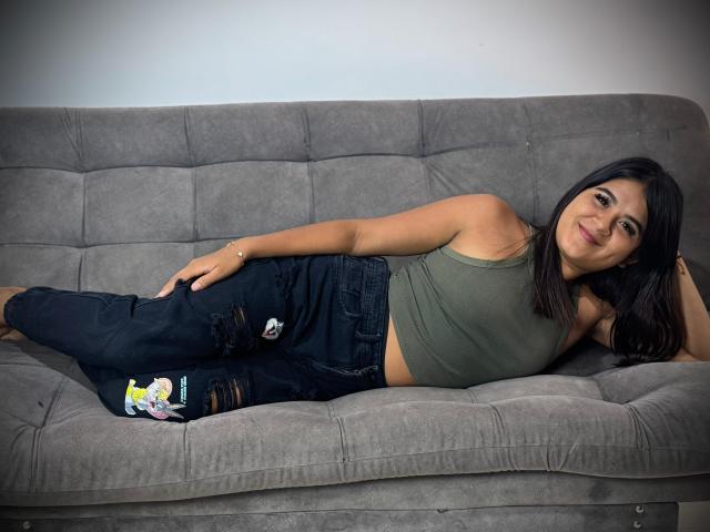 NaylaDiamond, lying on a grey sofa, wearing a green tank top and ripped blue jeans, with a small colorful sticker on her right thigh, smiling at the camera.