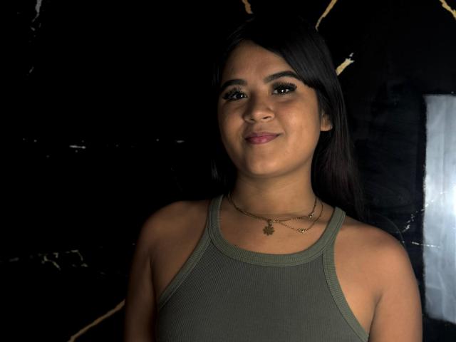 NaylaDiamond has tan skin, black hair, and a small smile. She's wearing a green tank top and two necklaces, with her tits slightly visible. Dark background.