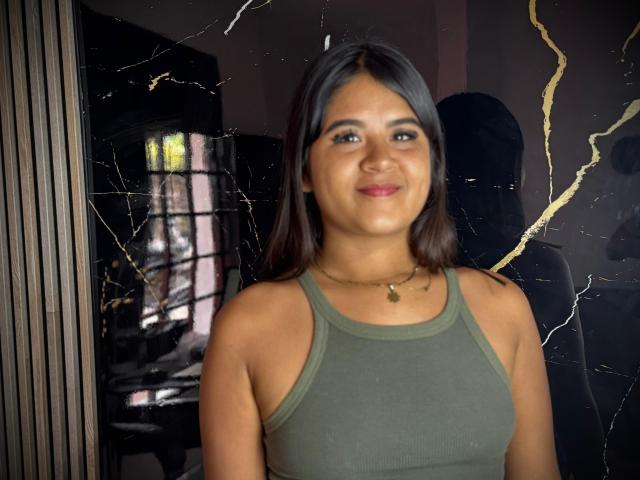 NaylaDiamond with medium brown skin and black hair, wearing a green tank top, smiling, and a gold necklace with a small pendant, standing in front of a cracked mirror.