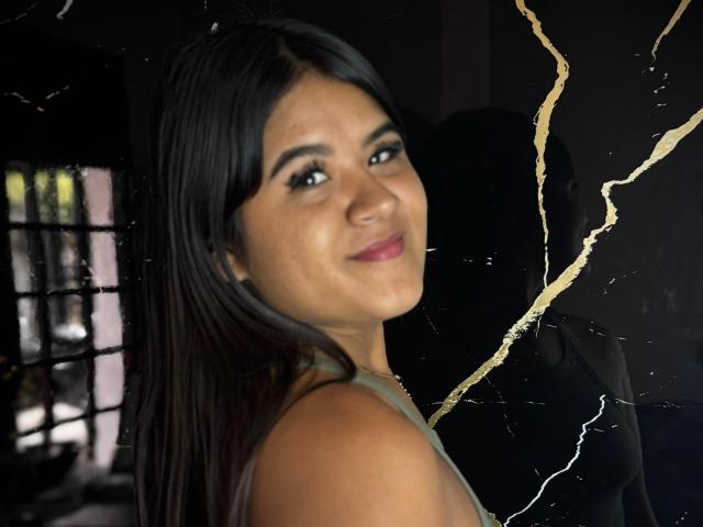 NaylaDiamond has medium brown skin, long black hair, and a slight smile. She's wearing a green top, with gold lines on a dark background, and has small, dark nipples.