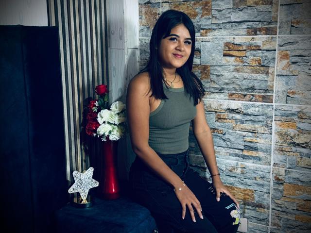 NaylaDiamond, brown-skinned, with black hair, green tank top, black ripped jeans, smiling, sitting on black chair, red vase with white and red flowers, and star decoration.