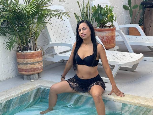 HazelDior with long black hair, small tits, wearing a black bikini top and lace skirt, sitting on pool edge, legs in water, white chairs and plants in background.