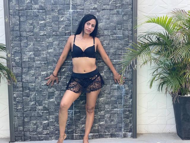 HazelDior with black lace bra and matching sheer shorts, standing against a gray stone wall, arms slightly out, looking at camera, dark hair, tan skin, small tattoo on right chest.