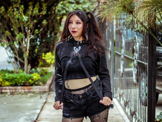 ScarlettPrincess with black leather crop top, black shorts, fishnet stockings, and puffy dark hair, standing in a sidewalk, showing her belly tattoo and holding straps.