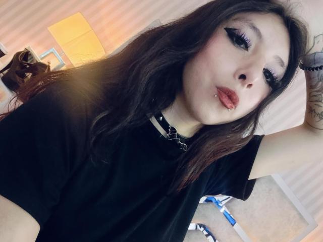ScarlettPrincess has long brown hair, black eyeliner, red lipstick, a lip piercing, and a black choker. She's wearing a black shirt, with her arm raised.