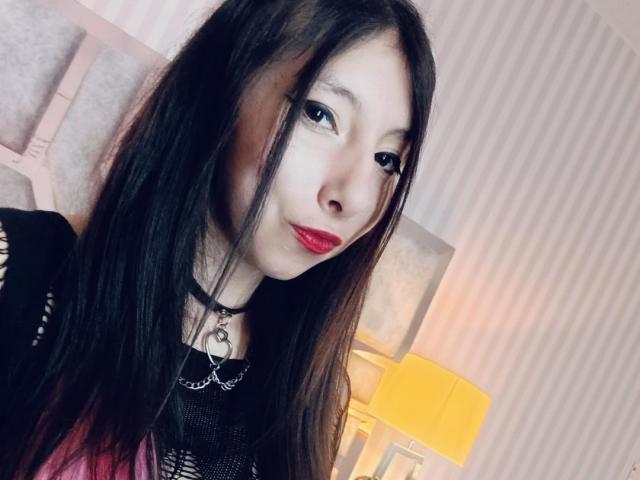 ScarlettPrincess has long black hair, red lipstick, and wears a black choker with a heart pendant, and a black mesh top. Her tits are small, and she looks sexy.