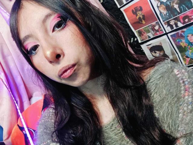ScarlettPrincess has long black hair, pink eye makeup, and full pink lips. She wears a green fuzzy top, with a soft pink curtain and colorful photos in the background.