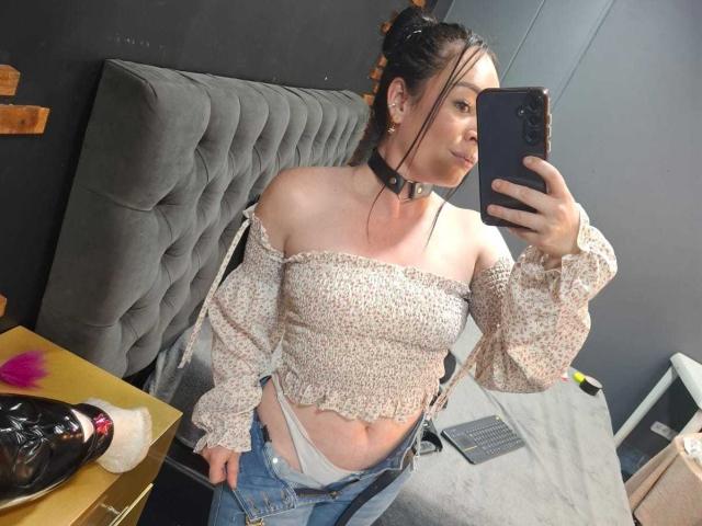 KristelRouse with a choker, off-shoulder floral top, and unbuttoned jeans showing white panties, taking a mirror selfie, her tits visible, dark hair in a bun, holding a phone.