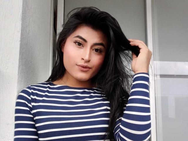 MichelleDenise has long black hair, wearing a blue and white striped shirt, looking at the camera with a slight smile, hand in her hair. She has a cute, sexy face.