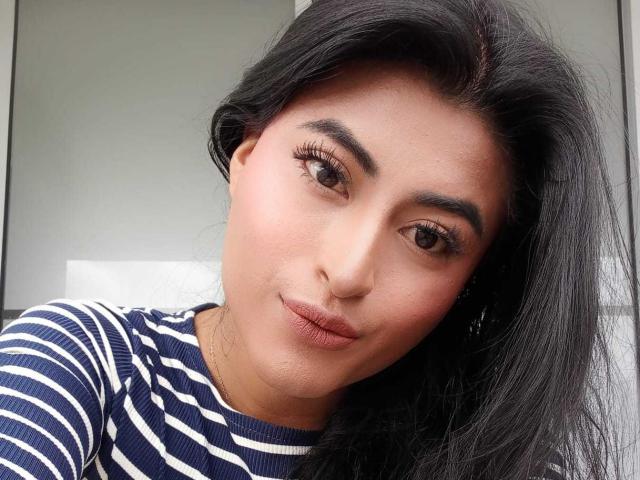 MichelleDenise has long black hair, brown eyes, and full lips with a slight smile. She's wearing a blue and white striped shirt, looking straight at the camera.