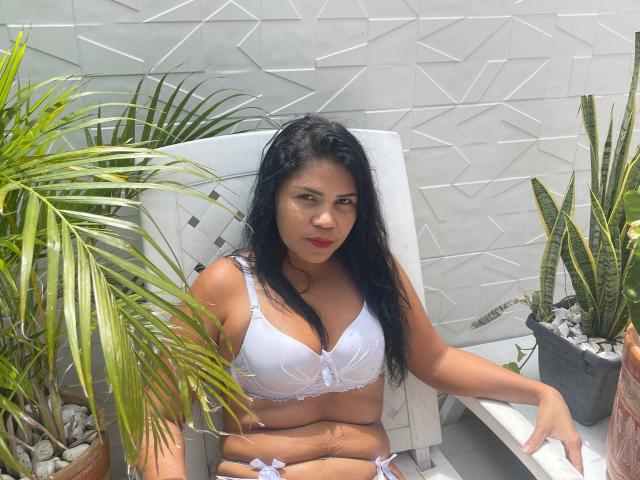 ZoeDior with long black hair, wearing a white bra and panties, sitting on a white chair, looking at the camera, surrounded by green plants, showing her tits and pussy.