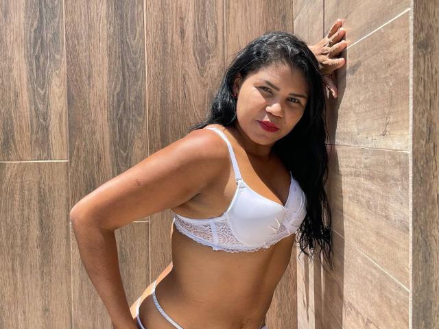 ZoeDior with dark hair, brown skin, and red lipstick, wearing a white lace bra and matching thong, leaning against wood-paneled wall, hands on hips, looking at camera.