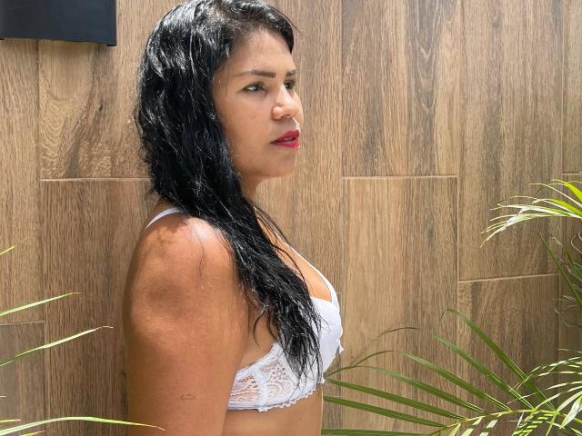 ZoeDior with wet black hair, red lipstick, and white lace bra, showing her small tits and brown skin, standing next to green plants. Wooden wall in the background.
