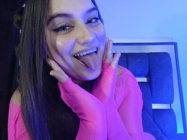 TaniaCooper69 with long dark hair, pink fishnet sleeves, and a playful expression, sticking out her tongue, hands on cheeks, blue lighting, and pink nail polish.