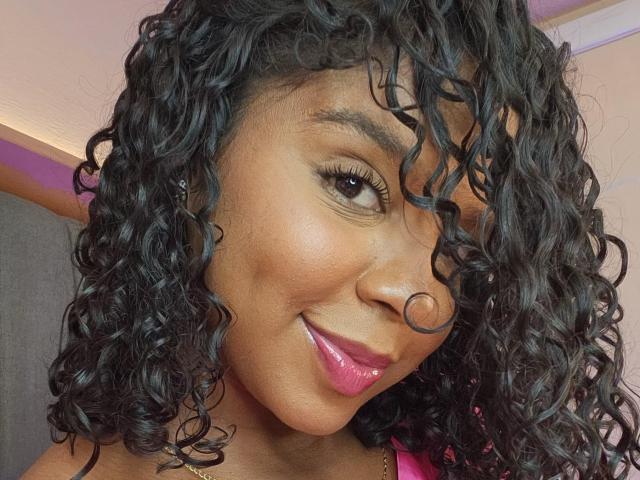 Das Gesicht von LisaMendez, lockiges, schwarzes Haar, dunkelbraune Haut, glänzender, rosafarbener Lippenstift, ein leichtes Lächeln, leicht gewelltes Haar, das ein Auge bedeckt, dezentes Make-up, sichtbare Wimpern, ein selbstbewusster Ausdruck, ein rosafarbener Akzent..
