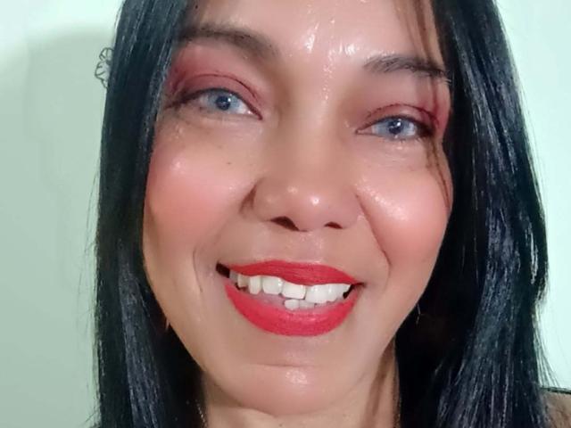 SashaBrownX tiene el cabello negro y brillante, ojos azules, sombra de ojos roja y un lápiz labial rojo intenso, con una ligera sonrisa que muestra dientes blancos, y un rostro húmedo y brillante..