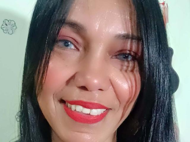 SashaBrownX has wet black hair, red lipstick, and a shiny face with water streaks. She's smiling with blue eyes and slight makeup. Her face is glistening.