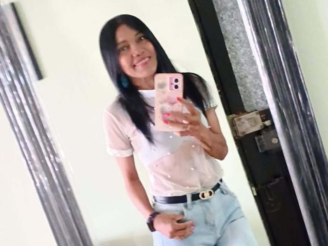 SashaBrownX, avec de longs cheveux noirs, une chemise blanche transparente, un jean bleu clair et une ceinture noire, tenant un téléphone rose, souriante, avec un reflet dans le miroir. Ses seins sont visibles à travers la chemise..