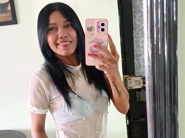 SashaBrownX, con cabello negro largo, sonriendo, sosteniendo un teléfono rosa, vistiendo una blusa blanca transparente con pequeños puntos blancos, mostrando sus pequeños pechos firmes, y uñas rojas..