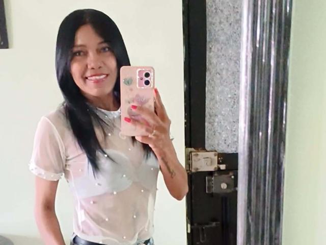 SashaBrownX, con cabello negro largo, sonriendo, vestida con una blusa blanca transparente que deja ver los pezones, sosteniendo un teléfono rosa, de pie frente a un espejo, mostrando un tatuaje en su brazo derecho..