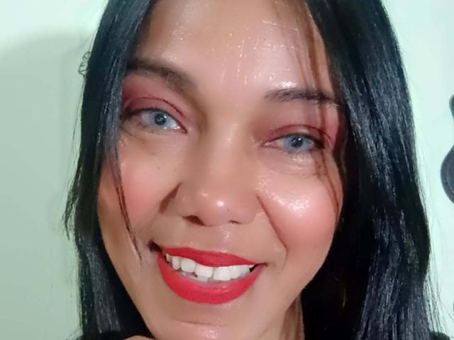 SashaBrownX tiene el cabello negro y brillante, ojos azules, lápiz labial rojo y sombra de ojos rosa. Está sonriendo con su rostro brillante y húmedo, y sus mejillas ligeramente sonrojadas. Sus senos no son visibles..