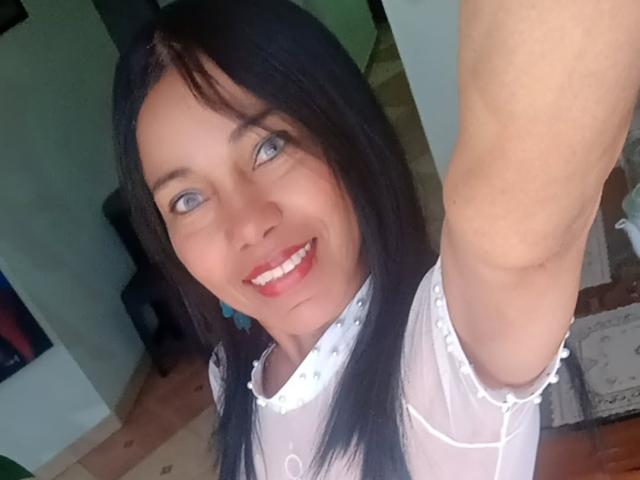 SashaBrownX a les cheveux noirs, les yeux bleus, et elle sourit avec du rouge à lèvres. Elle porte un haut blanc et transparent avec des décorations ressemblant à des perles. Son bras est levé pour un selfie..