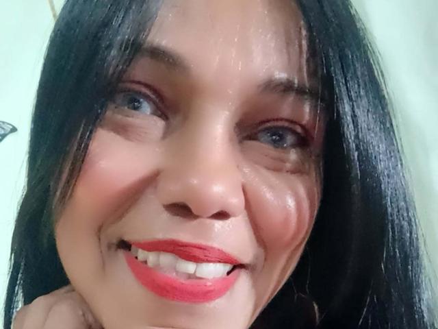 SashaBrownX hat glänzende, feuchte Haut, schwarzes, glattes Haar, blaue Augen, roten Lippenstift und ein leichtes Lächeln, das weiße Zähne zeigt. Ihr Gesicht glänzt vor Schweiß..