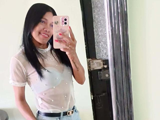 SashaBrownX with long black hair, wearing a sheer white top showing small, visible nipples, and light blue jeans, smiling while taking a mirror selfie with a pink phone.