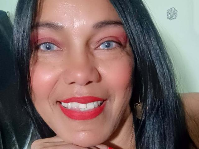 SashaBrownX tiene ojos azules brillantes, sombra de ojos roja y lápiz labial rojo, con cabello negro y lacio, y una sonrisa que muestra dientes blancos, luciendo linda y sexy con un ligero bronceado..