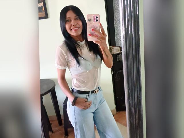 SashaBrownX, con i capelli neri, sorridente, indossa una camicetta bianca trasparente, abbottonata, e pantaloni a vita alta di colore azzurro chiaro, sta scattando un selfie allo specchio con un telefono rosa, mostrando il seno..
