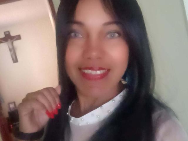 SashaBrownX a les cheveux noirs, les yeux bleus, du rouge à lèvres et un collier blanc orné de clous. Elle sourit, ses ongles sont rouges, et elle tient ses cheveux. Ses seins ne sont pas visibles..