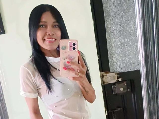 SashaBrownX, con cabello negro largo, sonriendo, sosteniendo un teléfono rosa, vistiendo una blusa blanca transparente, uñas rosas, pezones visibles, selfie frente al espejo, de pie en una habitación..