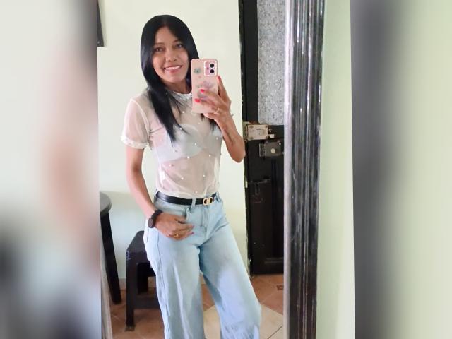 SashaBrownX with black hair, smiling, wearing a sheer white top with small studs, light blue high-waisted pants, taking a mirror selfie, holding a pink phone.