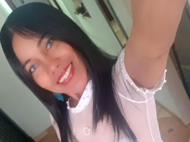 SashaBrownX, con cabello negro, ojos azules y lápiz labial rojo, sonriendo a la cámara. Lleva una blusa blanca y transparente con mangas fruncidas, que deja entrever parte de sus senos..