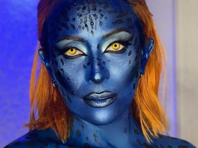 Sahrataylorr has blue face paint with black spots, yellow eyes, orange hair, and shiny blue lips. Her skin is blue, and she looks straight at the camera.